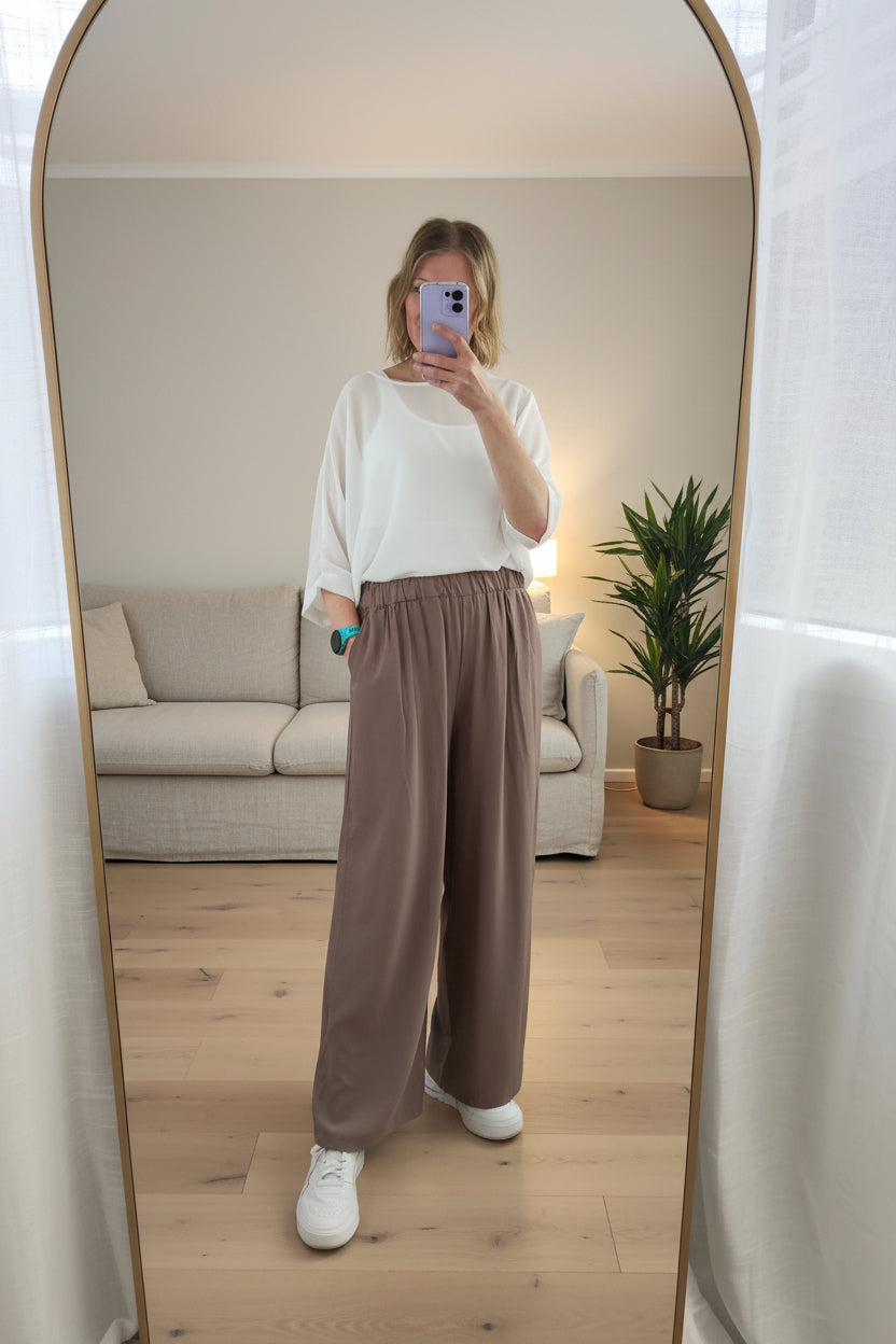 Person wearing a white top and brown pants taking a mirror selfie.
